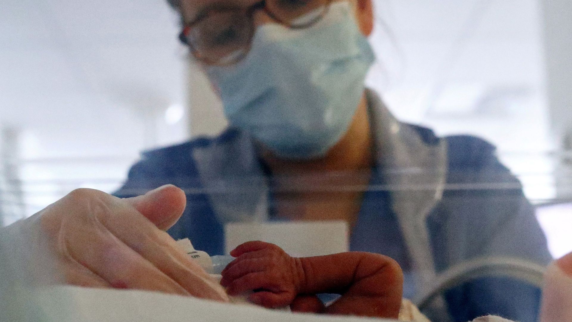 A neonatal nurse leans over a clear crib containing a baby, whose tiny arm can just be seen