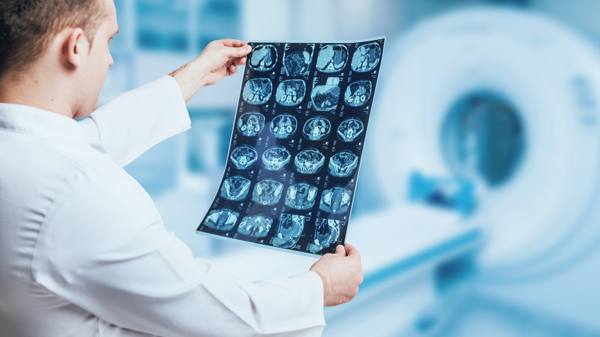 A doctor wearing a white coat and examining a printed sheet of MRI scan results next to an MRI machine