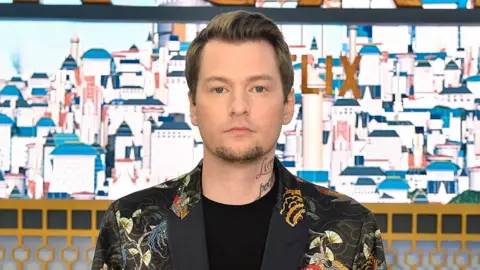 Getty Images A man with neatly cropped hair stands in front of a large picture of a town made up of tall, bright white buildings. It looks Meditteranean. He has handwriting tattooed on his neck and wears a black blazer with a traditional Japanese pattern featuring cranes and paper fans.