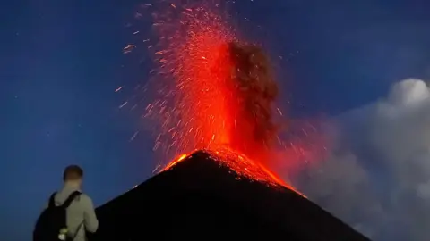 Louis Martlew Tourists look not far from an erupting volcano, which is spewing red and orange lava