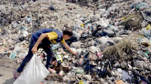 Mohamed searches through rubbish pile holding white bag