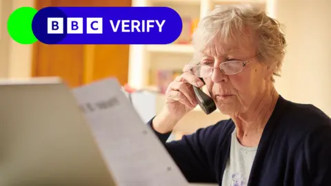 Getty Images A stock image of a grey-haired woman looking at a bill and her laptop while she is on the phone