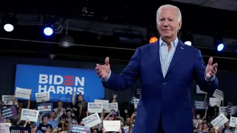 Reuters Biden in a blue blazer with hands out, in front of a crowd and large Biden Harris sign