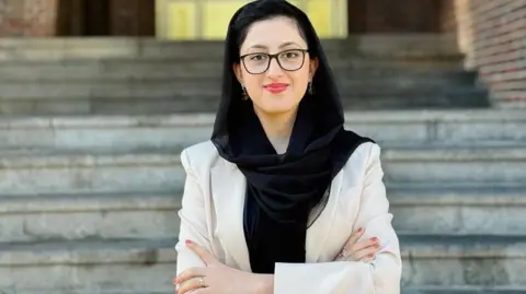 Parwana Ibrahimkhail Nijrabi  Spectacled Parwana Ibrahimkhail Nijrabi wearing a sandal-coloured coat and a black scarf over her head. 