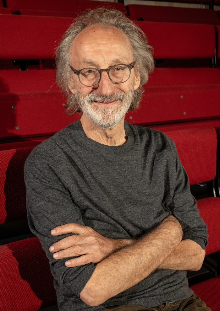 A man with grey hair, glasses and arms folded sits in a bank of red chairs