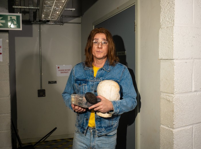 A man appears to be backstage. He has long hair, round glasses and is carrying a wig stand and make-up in a box