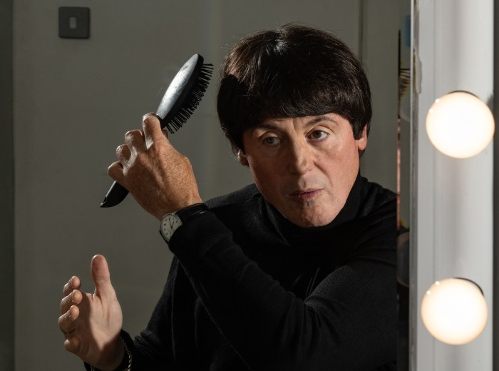 A man brushes his hair in a mirror with lights around it