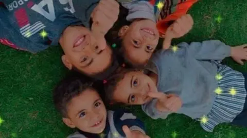 Handout Four children lay in circle on grass with their thumbs raised into the air. Some yellow stars, added via a camera filter, appear over the top of the image.