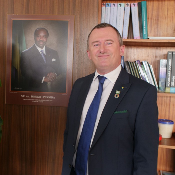 man in dark blue suit smiles at the camera, there is a photo of another man framed on the wall