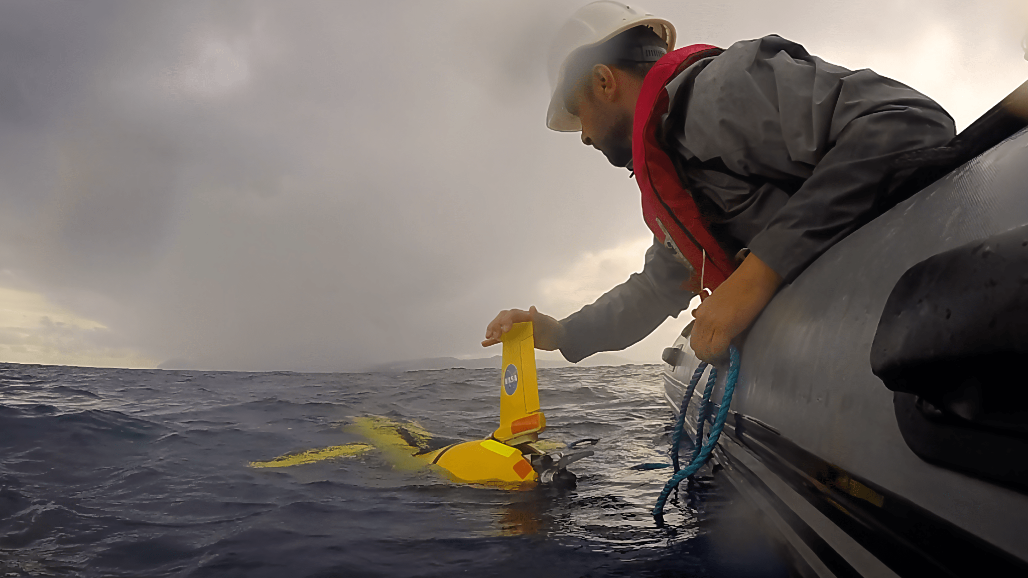 A hard-hat wearing scientist leans out of a zodiac boat to gently deploy an autonomous torpedo-shaped vehicle with wings, called a glider, into the ocean off the coast of Santorini, Greece.