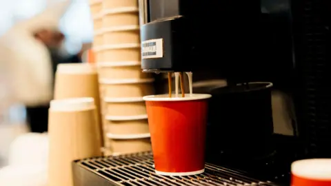 Getty Images A coffee machine dispensing milk and coffee into an orange paper cup. There is a tower of brown papers cups in the background.