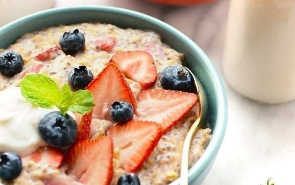 A bowl of creamy oatmeal is topped with sliced strawberries, blueberries, a dollop of yogurt, and a sprig of mint. The light blue bowl holds a spoon, resting against the sweet arrangement. The background features a light-colored surface with a partially visible container. MyFitnessPal Blog