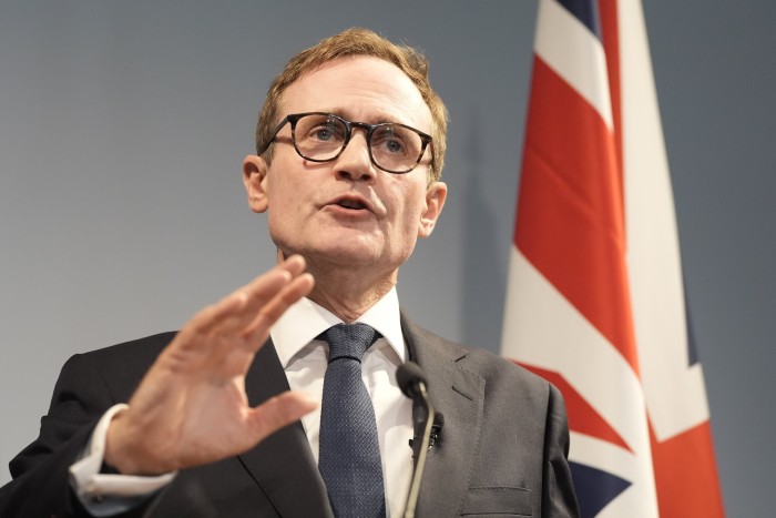 Tom Tugendhat, a Conservative party leadership candidate, is giving a speech in central London about delivering public services. In the image, he is seen  wearing a suit and tie, with a British flag in the background.