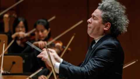 A man wearing a dark suit stands in front of an orchestra