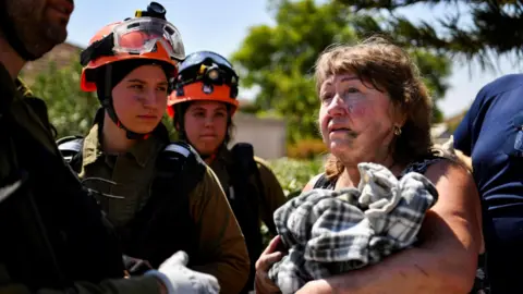 Reuters A woman speaks to Israeli first responders after her home was damaged by Hezbollah rocket fire in Katzrin, in the Israeli-occupied Golan Heights (21 August 2024)