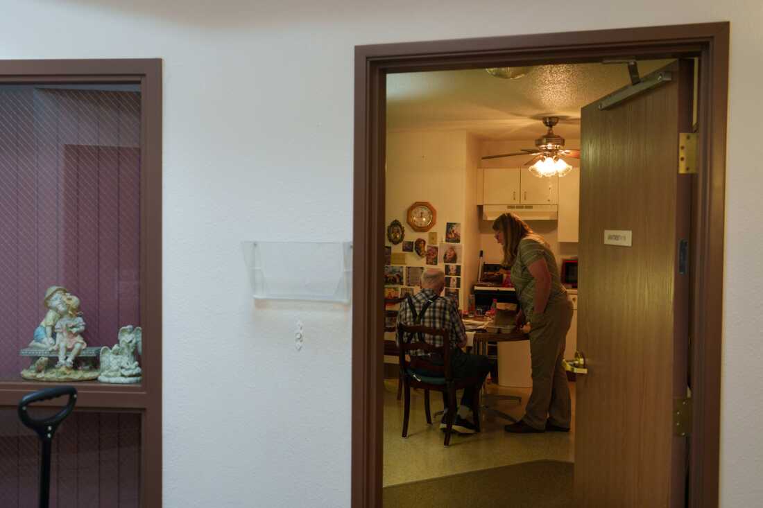 This photo shows Kyla Sanders delivering a meal to a resident at the Marian Manor senior apartments in Glen Ullin, N.D., on May 23. An older man wearing a plaid shirt and suspenders is seated at a kitchen table with his back to the camera. Sanders is standing next to the table with a hand near a brown paper bag. Kitchen cupboards and a stove are in the background.