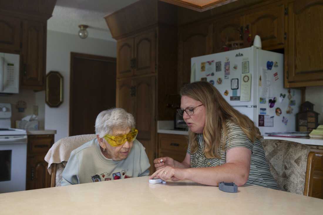 This photo shows Kyla Sanders helping 94-year-old Leona Staiger set up a medical alert device at her home in Hebron, N.D., on May 23. Both women are seated at a kitchen table, with Sanders on the right. Sanders, who has long hair and is wearing glasses and a horizontal-striped, short-sleeved shirt, is touching the device, which sits on the kitchen table. Staiger, who has short, silvery hair and is wearing a light-colored sweatshirt and glasses, is learning forward to look at the device. Kitchen cupboards, countertops and a fridge are in the background.