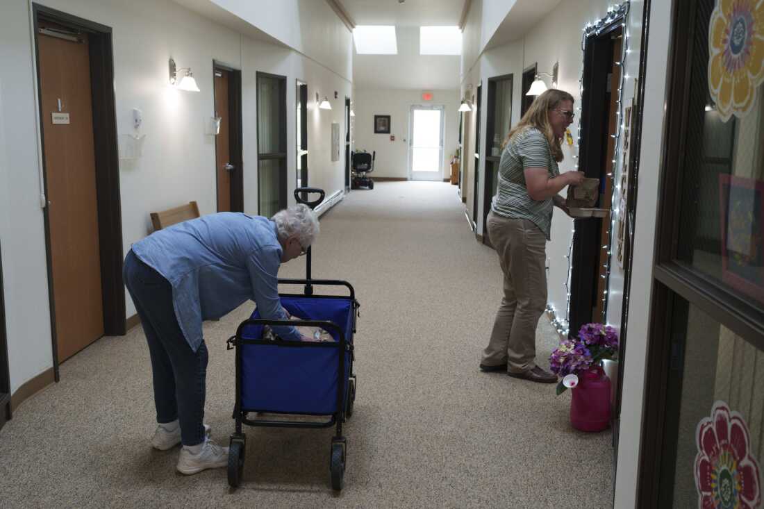 This photo shows Kyla Sanders helping to deliver meals to residents at the Marian Manor senior apartments in Glen Ullin, N.D., on May 23. Wearing khaki pants and a striped, short-sleeved shirt, she's standing in front of a doorway on the right side of a hallway and is holding a brown paper bag and a container of food. Also in the hallway is a silver-haired woman wearing blue pants and a blue shirt. She's bending down toward a blue wagon to retrieve a brown paper package from it.