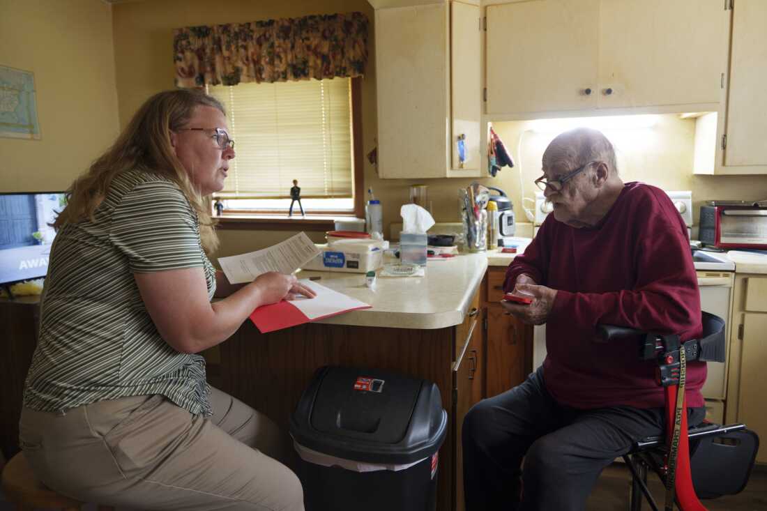 Kyla Sanders helps Don Fitterer, 81, fill out a health directive for local EMTs at his home in Glen Ullin, N.D., on May 23. Wearing dark pants and a dark red shirt, Fitterer sits near a kitchen counter on the right side of the photo. Sanders sits on the left side of the photo, wearing khaki pants and a short-sleeved shirt. She's holding papers in one hand; her other hand rests on an opened red folder on the countertop.