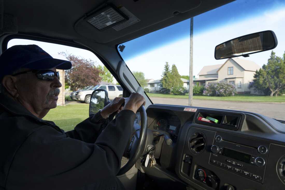 This photo shows Glen Ullin Ambulance Service EMT Dwight Kuntz driving the team's vehicle through Glen Ullin, N.D., on May 25. He is seated with his hands on the steering wheel and is wearing a dark shirt, sunglasses and a baseball cap. Through the vehicle's windshield, one can see a couple of houses.