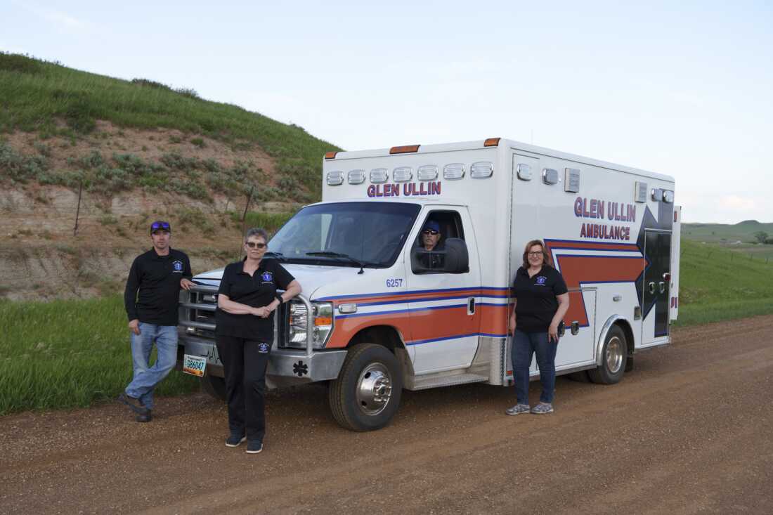 This photo shows Glen Ullin Ambulance Service EMTs Wade Kottre, Lori Kottre, Dwight Kuntz and Rita Wallin posing for a portrait on a county road in Glen Ullin, N.D., on May 25. Two of them are standing in front of an ambulance parked on the dirt road. One EMT sits in the ambulance's driver's seat, and the fourth EMT stands to the side of the ambulance. Behind them, the land slopes upward.