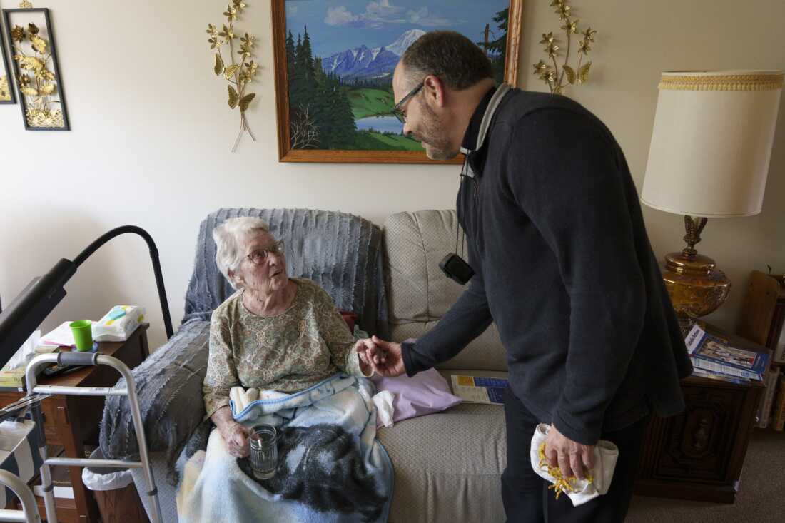 This photo shows the Rev. Gary Benz bidding farewell to Viola Weinhardt, 94, at her home in Glen Ullin, N.D., on May 24. Weinhardt is seated on a sofa with a blanket draped over her legs. A walker is on the left side of the photo. Benz is standing on the right side of the photo, with his hand outstretched and holding Weinhardt's hand.