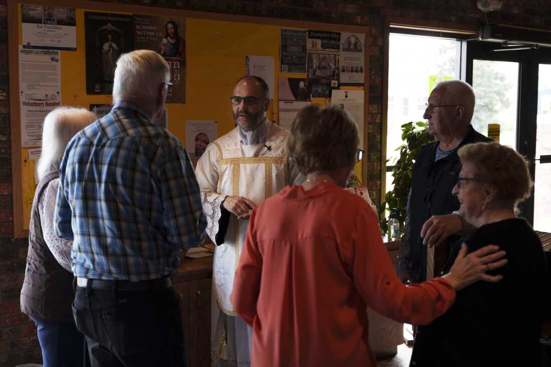 This photo shows the Rev. Gary Benz greeting parishioners after a Saturday afternoon Mass at the Sacred Heart Catholic Church in Glen Ullin, N.D., on May 24. Five older adults stand around him, with their backs to the camera. Benz is wearing a religious garment and is speaking to them. A bulletin board is behind him on the wall, displaying various flyers.