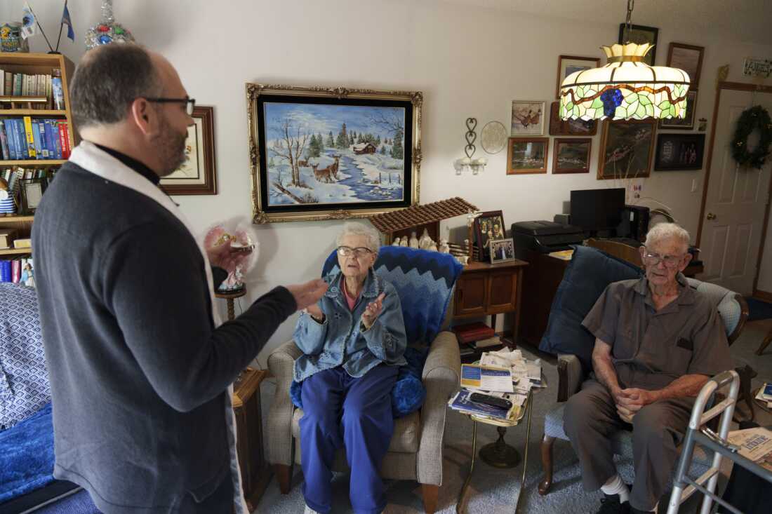 This photo shows the Rev. Gary Benz offering Holy Communion to Marianne and Jim Schaaf, both in their 90s, at their home in Glen Ullin, N.D., on May 24. Both Schaafs are seated in armchairs in their living room, and Benz is standing in front of them, facing them. Many pictures hang on the wall behind them.