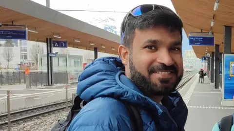 Syed Rafiq Syed Rafiq, a man with dark hair and a moustache and beard, smiles as he stands at a train station with a blue padded jacket on and sunglasses on the top of his head 