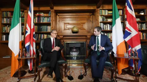 Charles McQuillan/PA Wire Sir Keir Starmer sitting with Simon Harris in library in Dublin's Farmleigh House, with British and Irish flags beside each of the two leaders