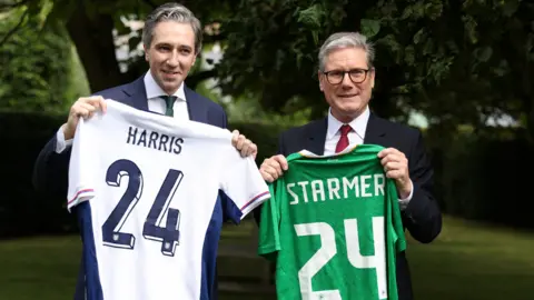 Reuters Taoiseach Simon Harris and Prime Minister Sir Keir Starmer holding up football jerseys with their surnames on the back of each shirt, along with the number 24. 