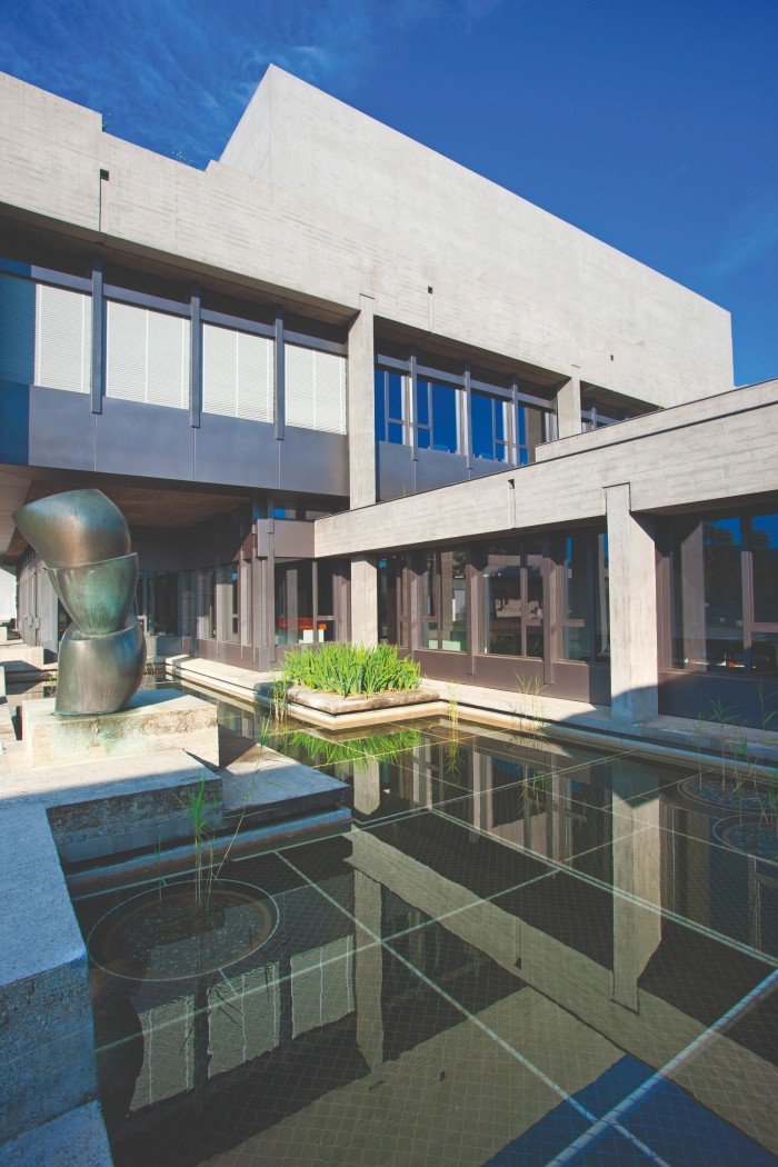 A modern architectural building with a concrete exterior and large windows, featuring a reflective water pond with green plants and a bronze abstract sculpture in the courtyard