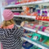 A girl with her back to the camera stands in a store, facing shelves of health and beauty products on display for sale. The girl appears to be the size of a tween or young teen and is wearing a black and white patterned coat and a pink winter cap.