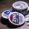 This photo shows two round, puck-shaped containers of Zyn nicotine pouches stacked together. The lid has been removed from the top container, and small white pouches of powdered nicotine are inside.