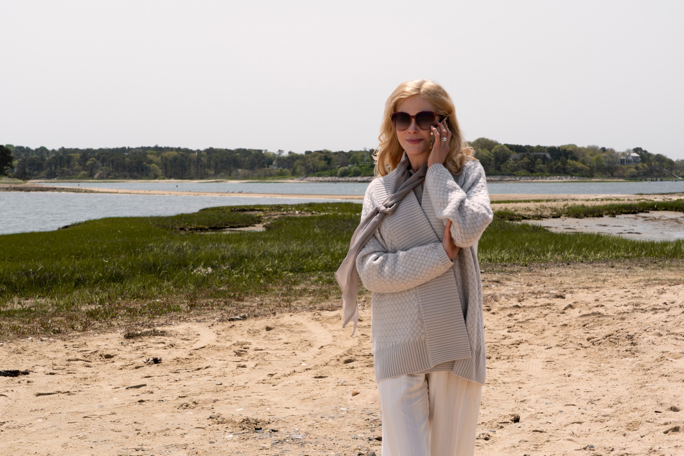 Nicole Kidman in sunglasses and a sweater holding a phone to her ear while walking along a coastal inlet.