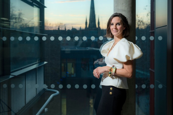 Alex Mahon in white blouse and black skirt stands in a glass elevator with view of city at dusk behind her