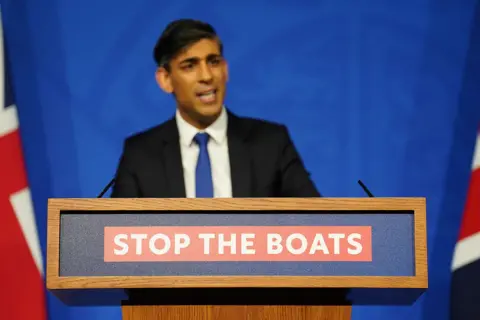 Reuters Rishi Sunak standing behind a podium with the slogan STOP THE BOATS emblazoned on it. There are Union flags on either side of him and a blue background.