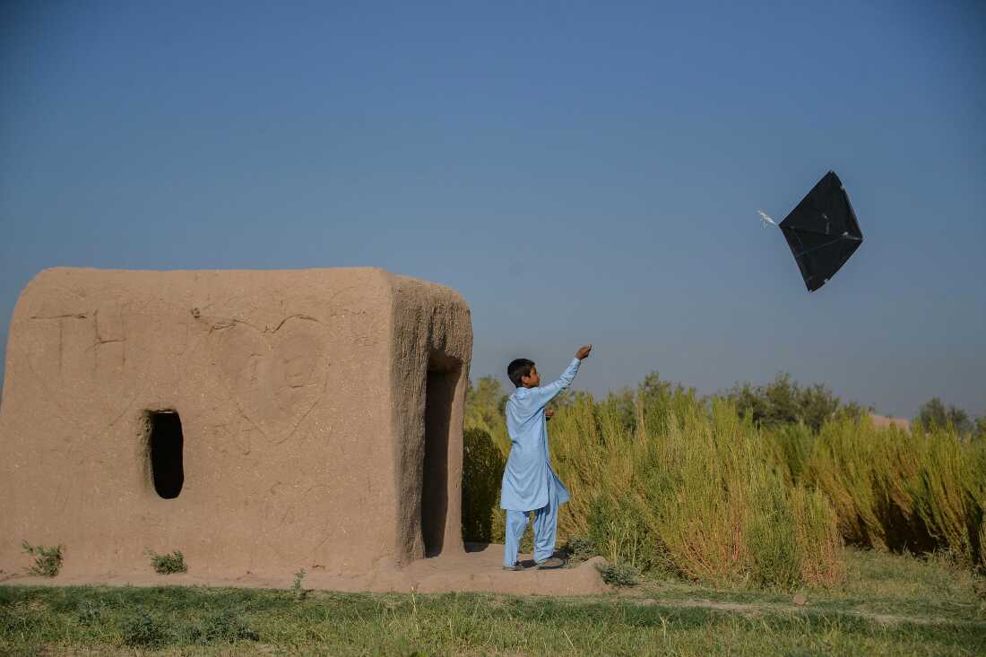 An Afghan boy wearing light blue clothes flies a kite while standing next to an earthen structure at the edge of an open field with high grasses.