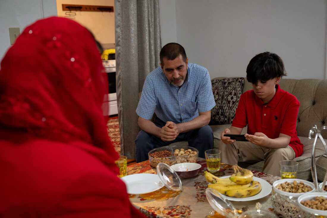 Mujib Ur Rahmani plays a video game on a phone in his living room in Lewiston, Maine, while his parents Mohammad Rahmani, center, and Khadija Rahmani, left, talk on Sunday, June 23, 2024. (Raquel C. Zaldívar/New England News Collaborative)