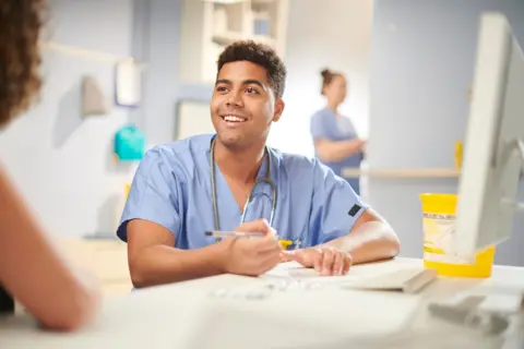 Getty Images A member of hospital staff with a stethoscope sitting at a computer in a clinical setting and holding a pen 