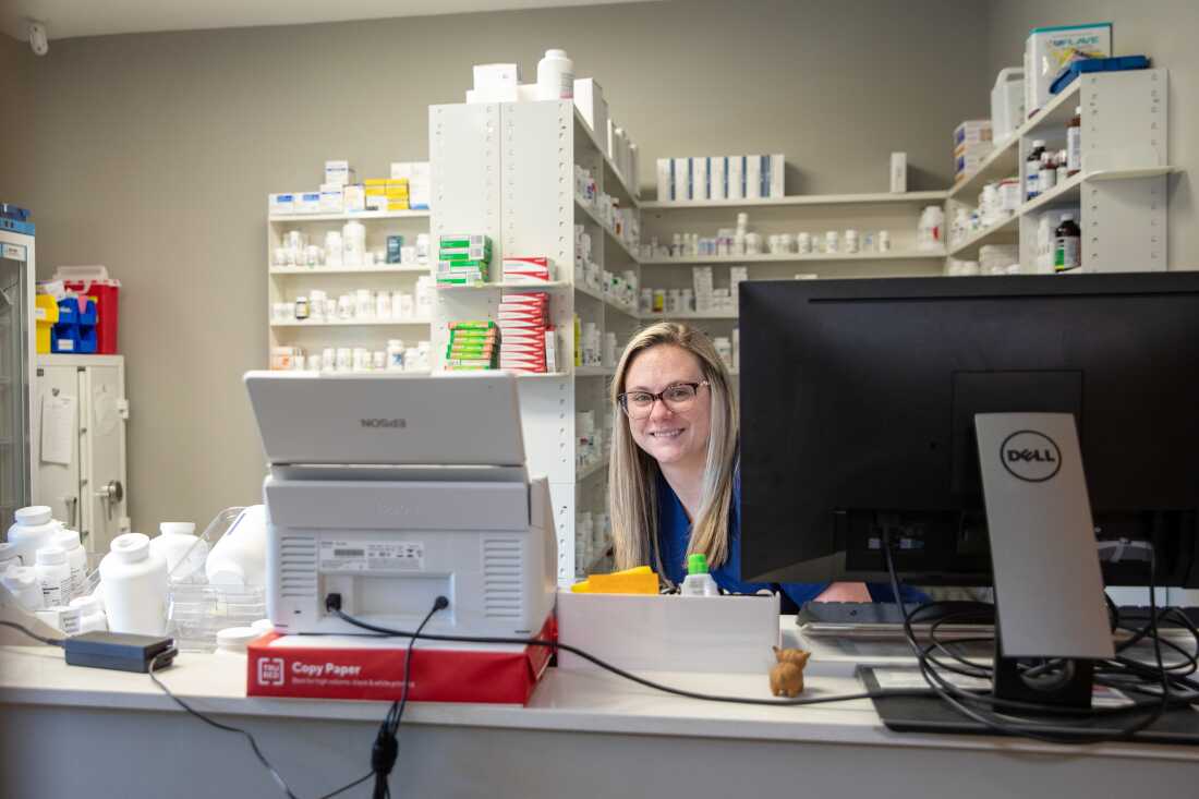 Brooke Williams works at the pharmacy counter at the PhiferCares Clinic outside Tuscaloosa, Alabama. At no cost, Phifer employees and their families can visit and get basic primary care, including prescription drugs. A smiling woman with blond hair sits behind a counter and a computer; shelves filled with medicines are in the background.