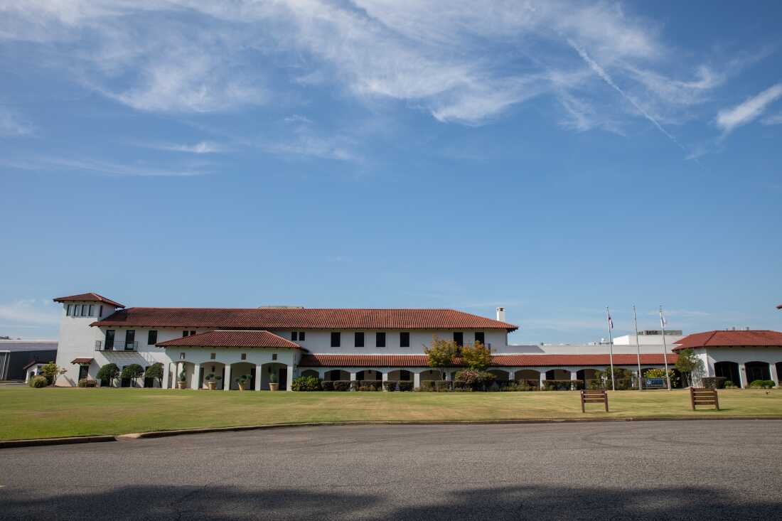 The exterior of the Phifer offices and factory outside Tuscaloosa, Alabama. The building is a sprawling, elegant white structure with a colanade and a red-tile roof. The photo shows a blue sky with wispy clouds and an expanse of green lawn.  