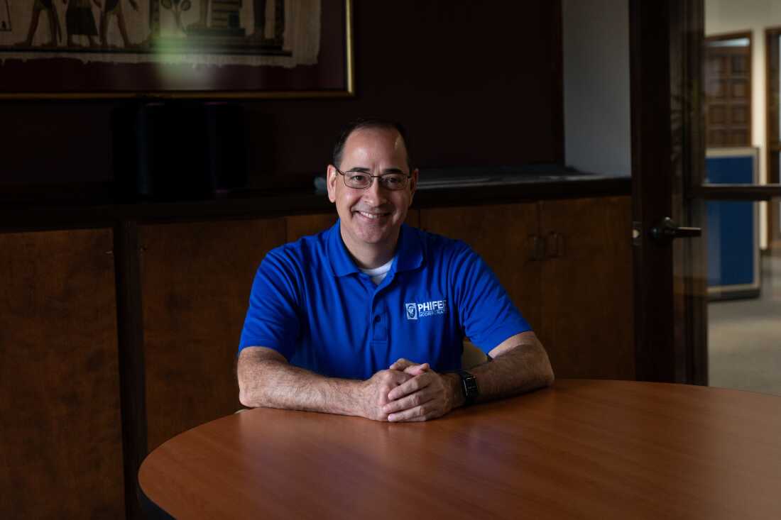 Russell DuBose, vice president of human resources at Phifer, says, “We don’t want you to spend money on health care.” He is a man with dark hair and glasses, sitting with his hands clasped together on a conference table. He is wearing a blue company golf shirt. 