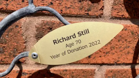 A gold-coloured piece of metal in the shape of a leaf is attached to a wavy piece of grey-coloured metal, with a redbrick wall behind it. The inscription in black writing on the leaf says: "Richard Still, Age 70, Year of Donation 2022."