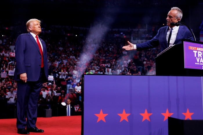 A man in a suit gives a speech and gestures towards former US president Donald Trump in a packed hall