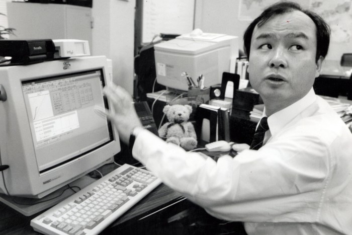 A man in shirt and tie sits at a desk in front of a computer screen. He is turning round to speak to someone behind him. On the desk is a small teddy