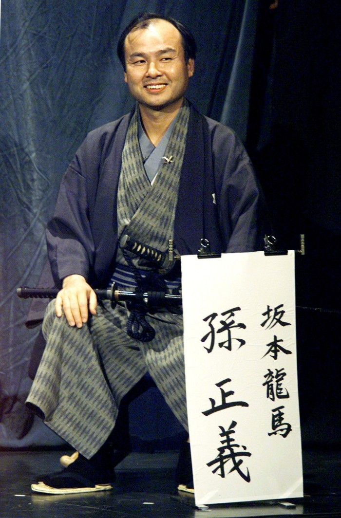 A man, smiling, is on stage, dressed in traditional samurai dress. He crouches down, his sword on his legs. In front of him is a sign with Japanese lettering