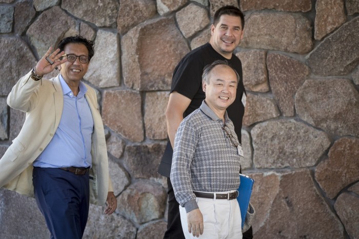 Three men, smiling, walk past a rustic-style wall. All are casually dressed. One of them, Misra, is waving in greeting