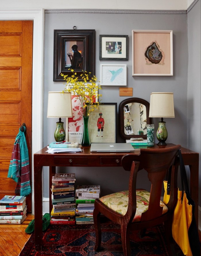 A room with a desk and table on which there is a laptop, pictures and two lamps, with pictures on the wall and piles of books on the floor