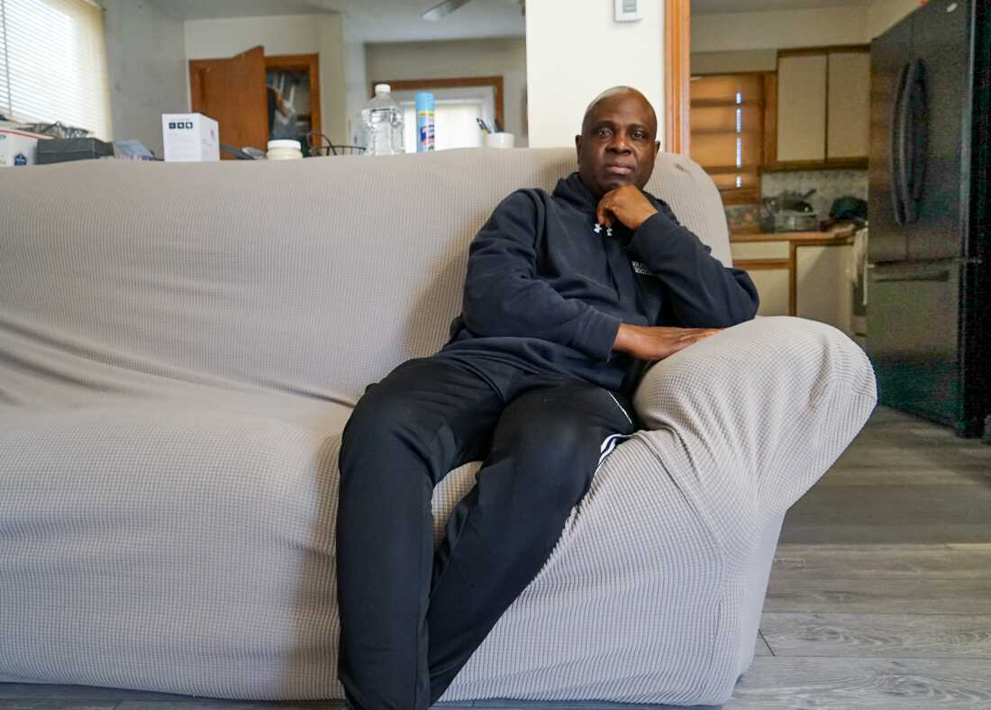 Nursing assistant Kwesi Ablordeppey sits on a grey couch wearing dark clothing. 